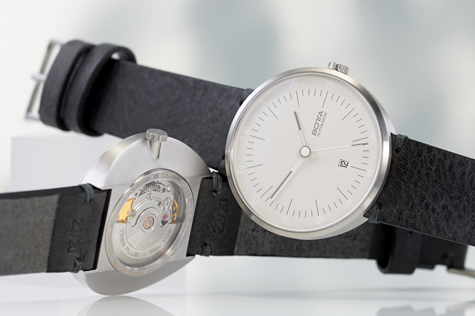 Abgebildet ist die TRES Automatik von BOTTA Design, eine moderne Bauhaus-inspirierte Designuhr. Das weiße Zifferblatt mit schlichten Indizes und Datum bei 6 Uhr unterstreicht die klare Formsprache. Durch den transparenten Gehäuseboden ist das Swiss-Made Sellita SW200-1 Automatikwerk sichtbar. Diese Automatikuhr aus Titan steht für Präzision und Zeitlosigkeit – ideal für alle, die eine besondere Armbanduhr beziehungsweise eine elegante Designuhr kaufen oder bestellen möchten.